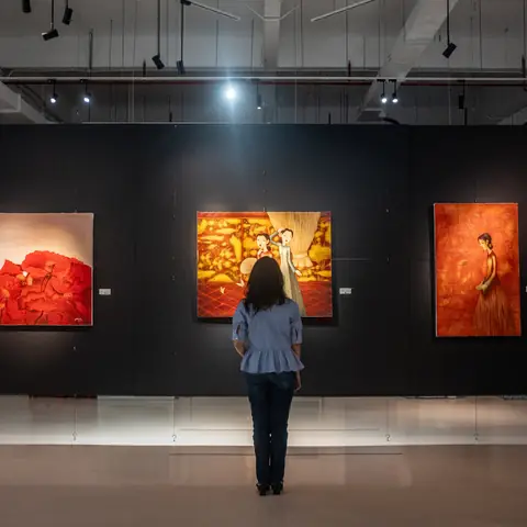 A Person Stands In A Gallery Space With Dark Walls Where Three Paintings With Red And Orange Colors Are Mounted